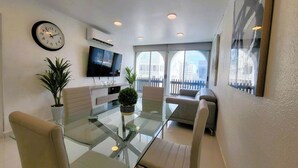 A modern living room with a glass dining table, comfortable seating, a flat-screen TV, and a balcony with a view.