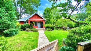 A red house with a front porch, surrounded by a well-maintained lawn and various shrubs. - Deluxe Downtown Bungalow - fenced in yard, pet friendly. (Brevard)