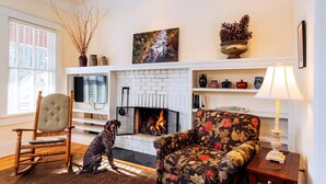 A living room with a fireplace, a dog, a floral armchair, and a wooden rocking chair. - Deluxe Downtown Bungalow - fenced in yard, pet friendly. (Brevard)