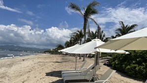 Ein Strand mit weißen Liegestühlen und Sonnenschirmen, Palmen und Blick auf den Ozean.