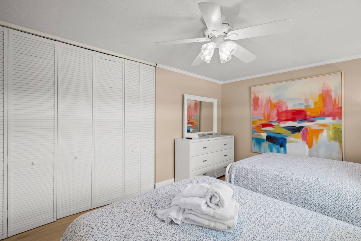 A bedroom with two beds, a white dresser, a ceiling fan, and a large abstract painting on the wall.