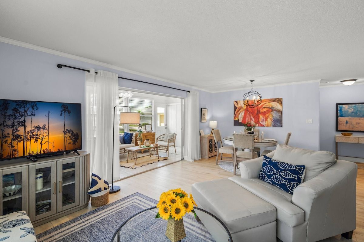 A modern living room with a flat-screen TV, a sofa, a coffee table with sunflowers, and a dining area in the background.