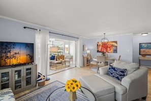 Ein modernes Wohnzimmer mit einem Flachbildfernseher, einer Couch, einem Couchtisch mit Sonnenblumen und einem Essbereich im Hintergrund.