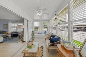 A spacious living area with a ceiling fan, wicker furniture, and a view through large windows.