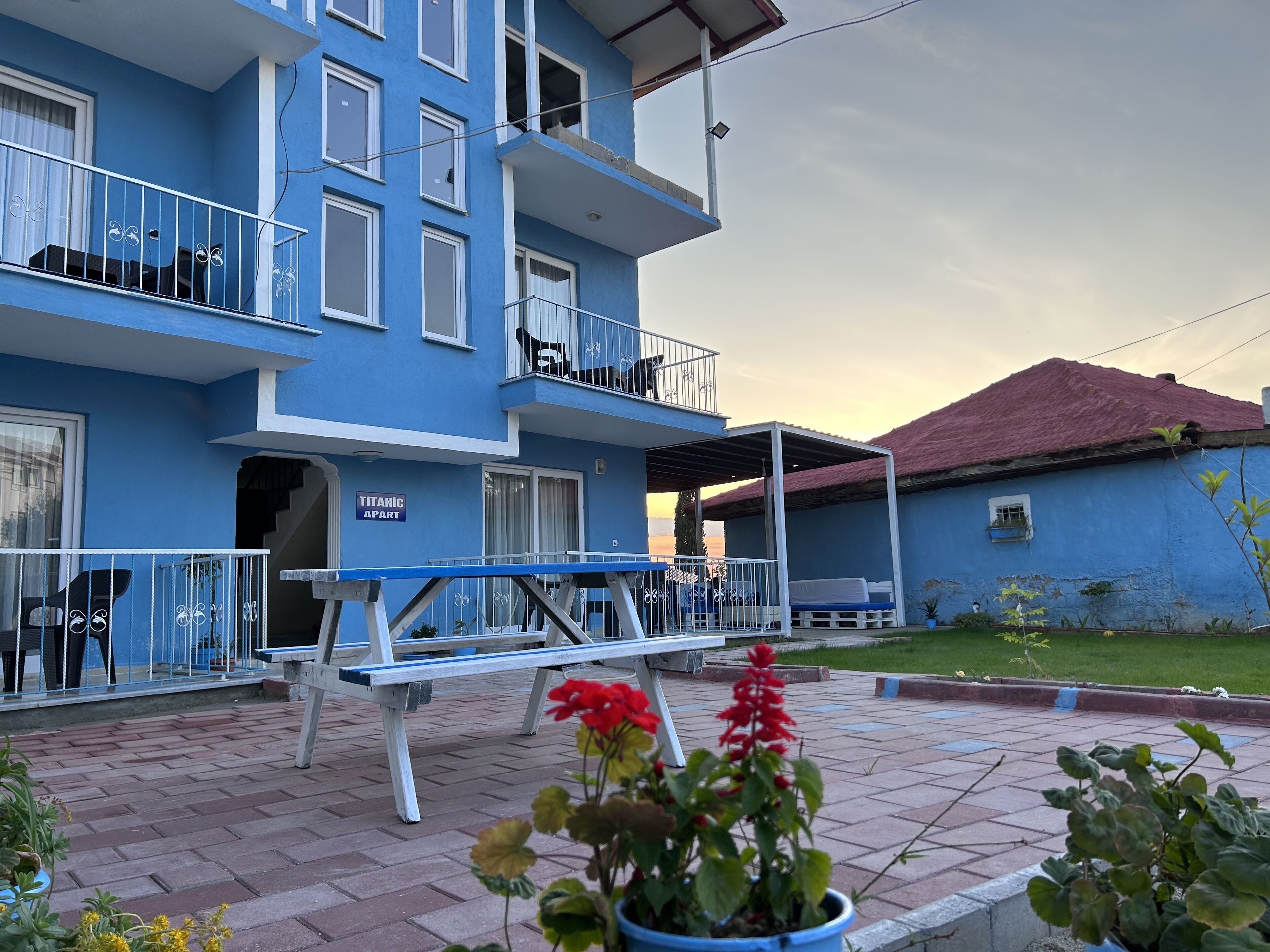 A blue building with balconies, a sign reading "TITANIC APART," and a patio area with a bench and potted plants.