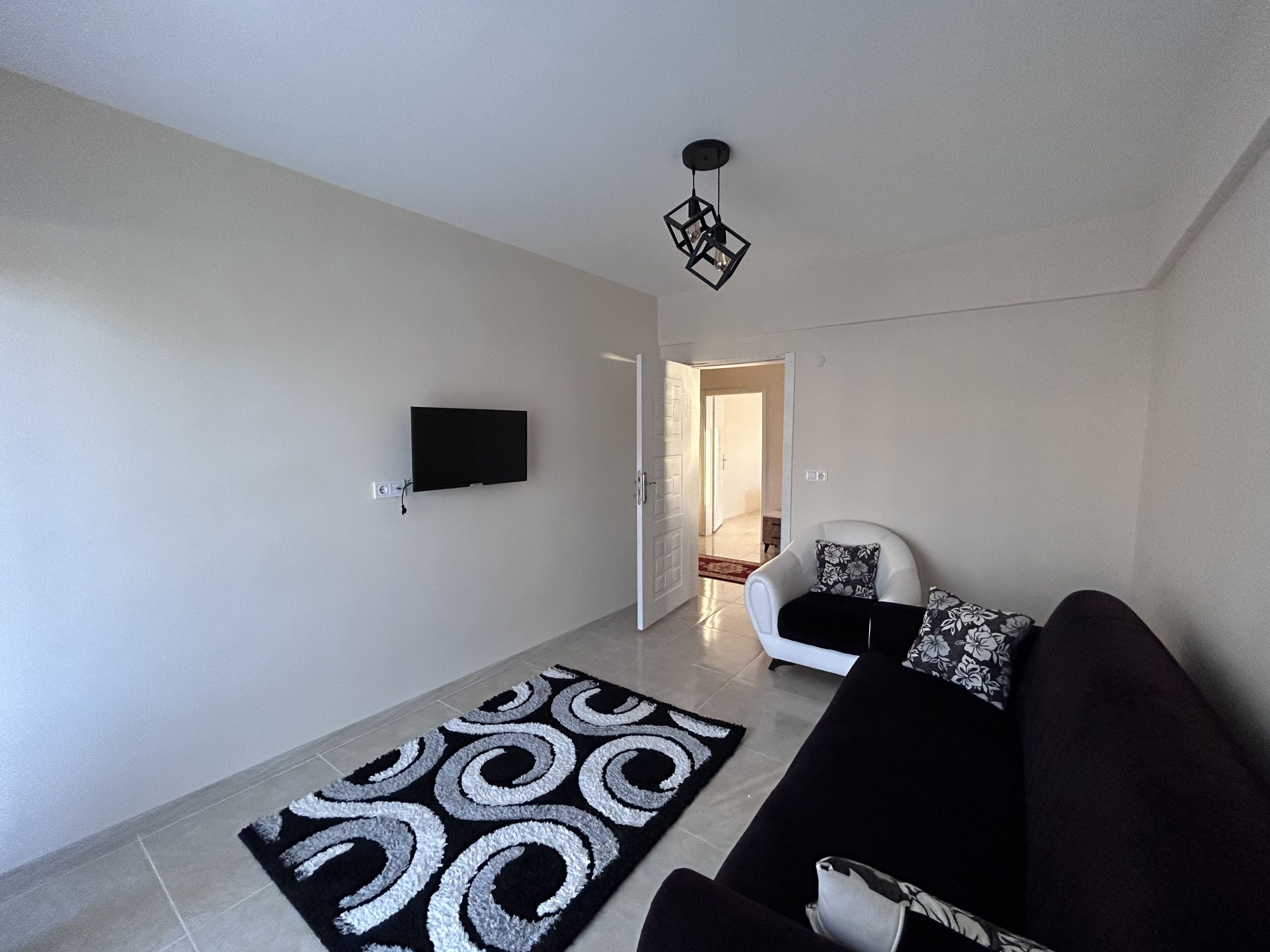 Un salon moderne avec un téléviseur à écran plat fixé au mur, un tapis à motifs noir et blanc, un canapé noir agrémenté de coussins à fleurs et un luminaire suspendu noir.