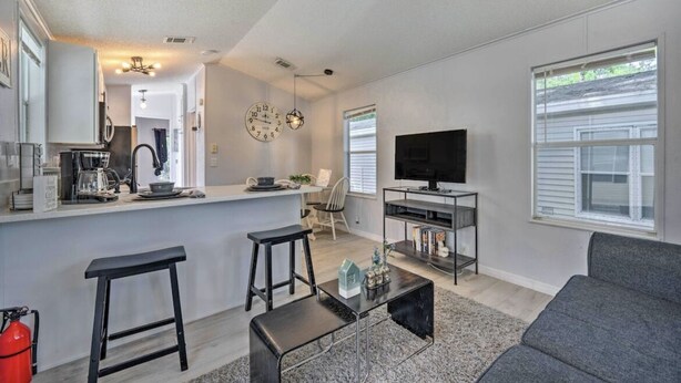 A modern living room with a grey sofa, a coffee table, a TV stand, and a kitchen area with a bar. - Enchanted Cottage with Pool and Mini-Golf Near Disney (Kissimmee)