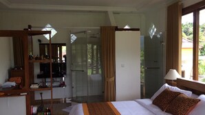A bedroom with a bed, a desk, a mirror, a shower, and a glass door leading to a balcony.