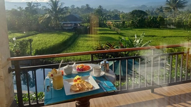 A balcony with a table set for breakfast, overlooking a lush green landscape with rice fields and palm trees, and a mountain range in the distance.