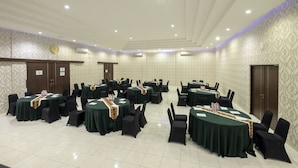 A conference room with multiple round tables, black chairs, and green tablecloths. There are air conditioning units on the walls, a clock, and a door on the left side. - Assalaam Syariah Hotel (Surakarta)