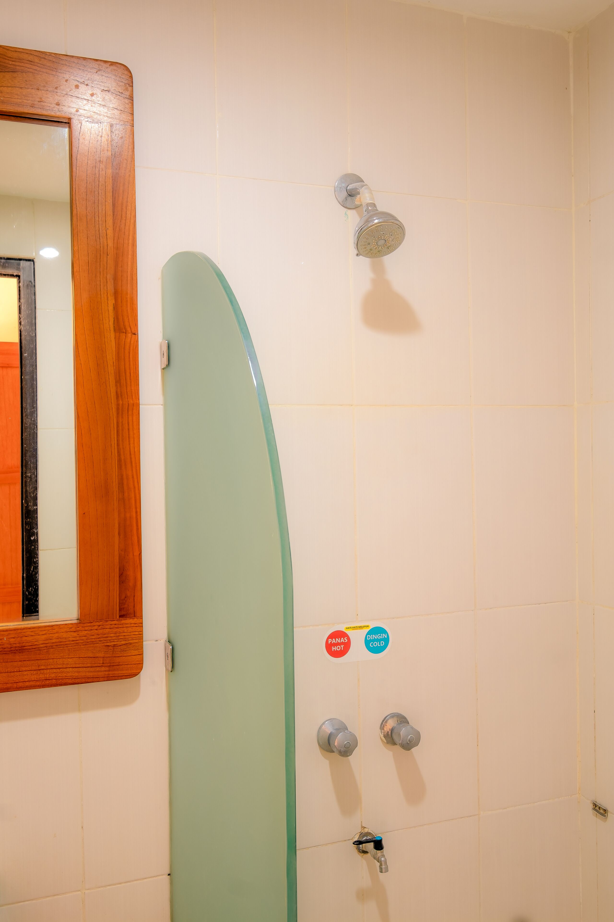 Une douche avec une porte en verre, un miroir encadré en bois et deux boutons de réglage de la température portant les inscriptions « PANAS HOT » et « DINGIN COLD ».