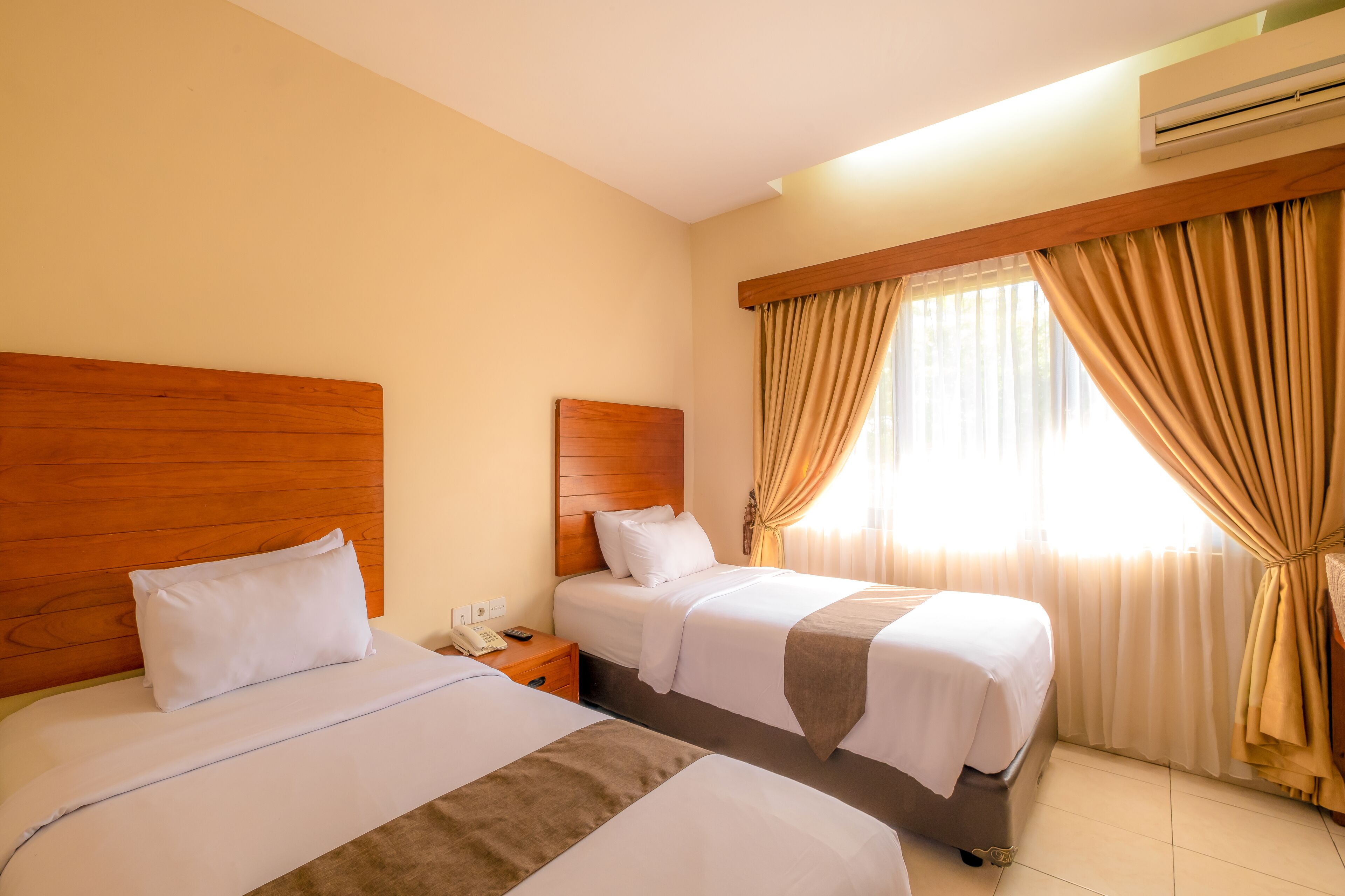 Habitación de hotel con dos camas, cabeceros de madera, paredes color crema y una ventana con cortinas.