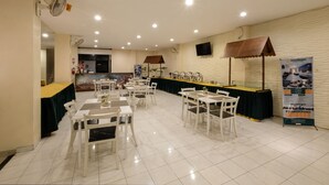 A spacious dining area with white tables and chairs, a buffet counter, and a wall-mounted TV. - Assalaam Syariah Hotel (Surakarta)