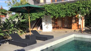 A swimming pool with lounge chairs and an umbrella, surrounded by greenery and a building with a staircase.
