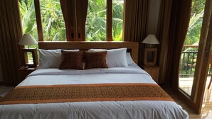 A large bed with white linens and a patterned throw blanket, flanked by two nightstands with lamps, in a room with large windows and curtains.