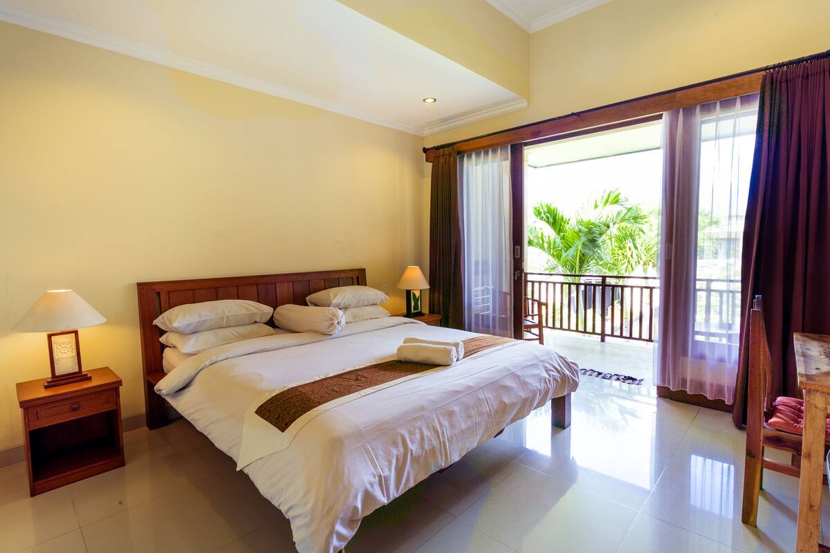 Habitación de hotel con una cama grande, dos mesitas de noche, un buró, un balcón con cortinas y vistas al verdor exterior.