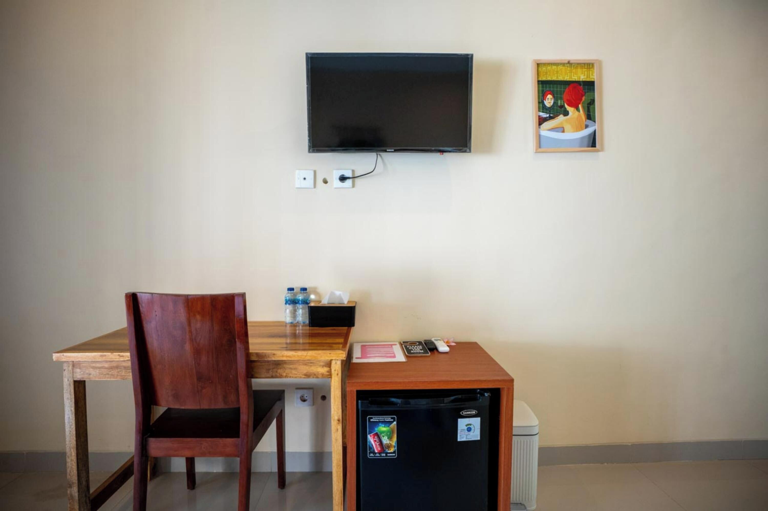A small room with a wooden desk, a chair, a television mounted on the wall, a refrigerator, and a framed picture.
