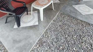 Ein überdachter Sitzbereich mit zwei Plastikstühlen und einem kleinen Tisch, eine Tür mit grünem Rahmen und eine rote Außenwand.