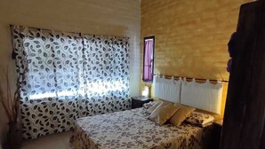 Ein Schlafzimmer mit einem Bett, einem Deckenventilator, einem Fenster mit gemusterten Vorhängen und einem Nachttisch mit Lampe.