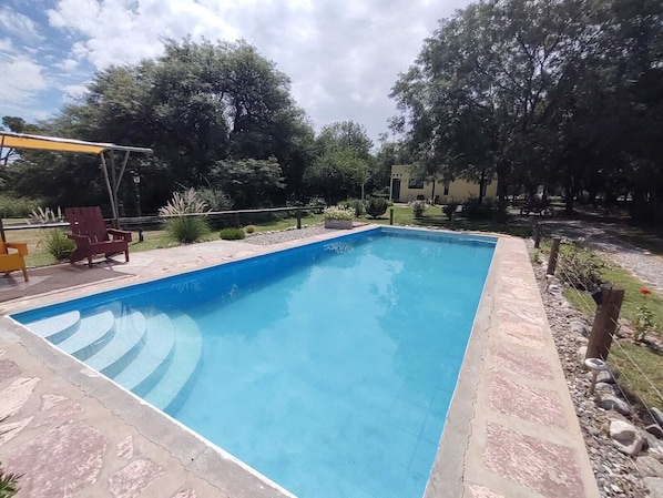 A rectangular swimming pool with a stone border, surrounded by a patio with chairs and a pergola.