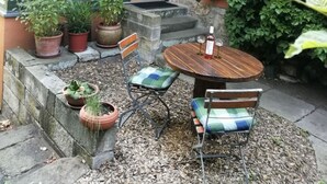Een terras met een houten tafel en stoelen, omgeven door plantenpotten en een stenen bank.