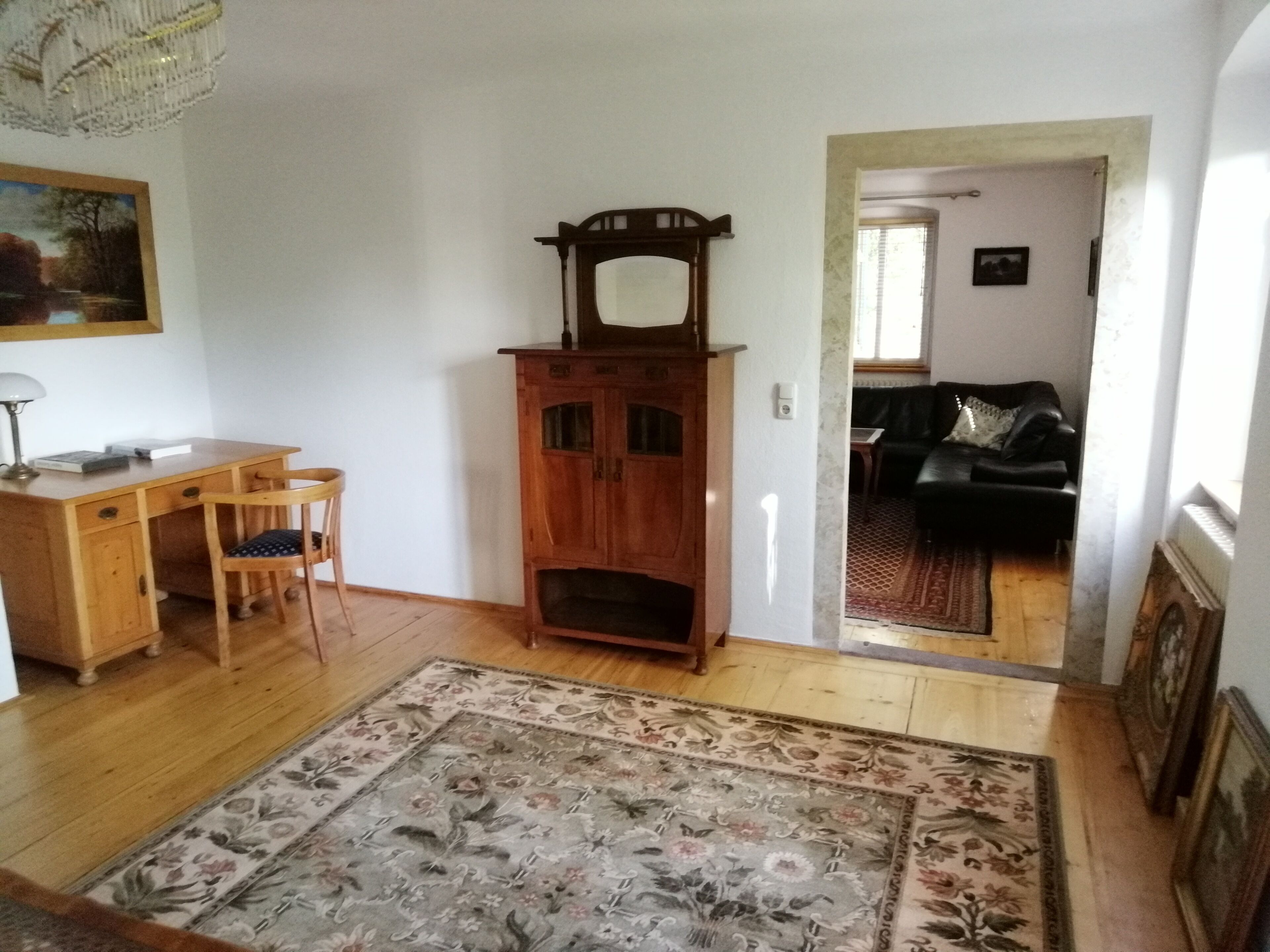Ein Zimmer mit einem Holztisch, einem Stuhl, einem Holzschrank mit Spiegel, einem Blumenteppich, einem Sofa und einem gerahmten Bild an der Wand.