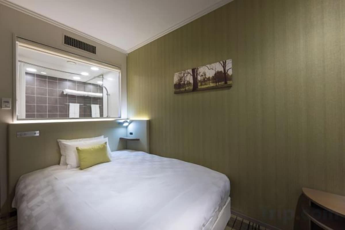Een hotelkamer met een bed, een nachtkastje, een wandlamp, een raam met uitzicht en een schilderij aan de muur.