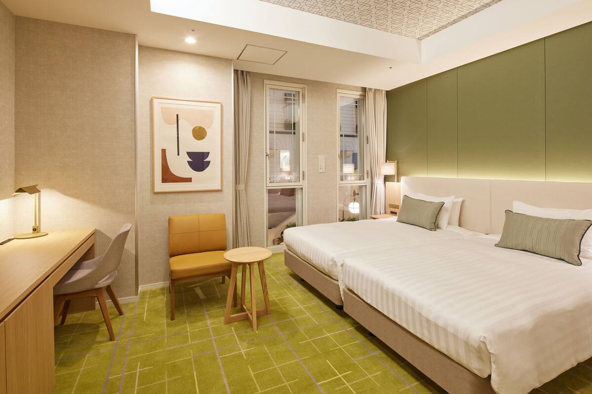 A hotel room with a bed, a desk, a chair, a small stool, a window with curtains, and a wall-mounted lamp.