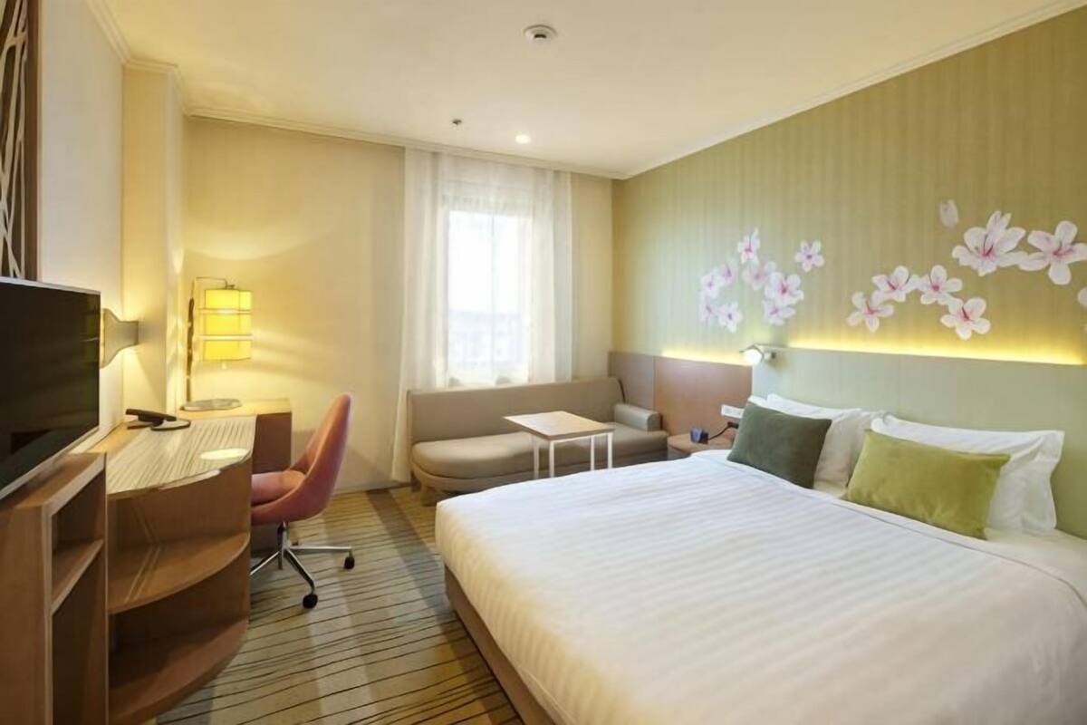 A hotel room with a bed, a desk with a chair, a sofa, a TV, and a window with curtains.