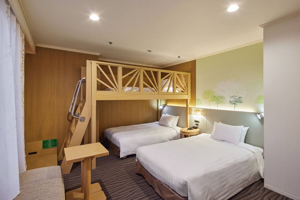 A hotel room with two beds, a wooden bunk bed, a small table, and a nightstand with a lamp.
