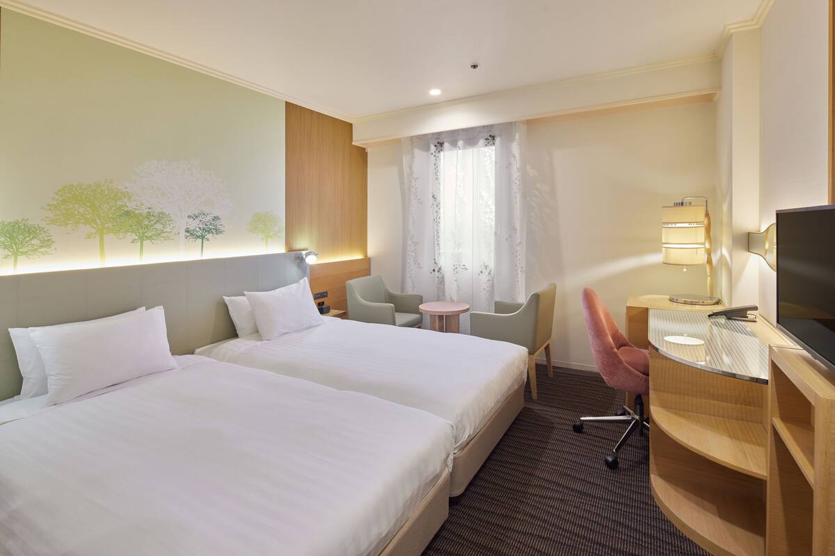 A hotel room with two beds, a desk, a chair, a TV, and a wall with a tree mural.