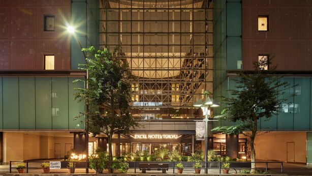 Un hotel con un amplio frente de vidrio, una entrada destacada con un letrero, y un espacio exterior iluminado con plantas en macetas.