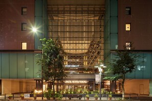 A hotel with a large glass facade, a prominent entrance with a sign, and a well-lit outdoor area with potted plants.
