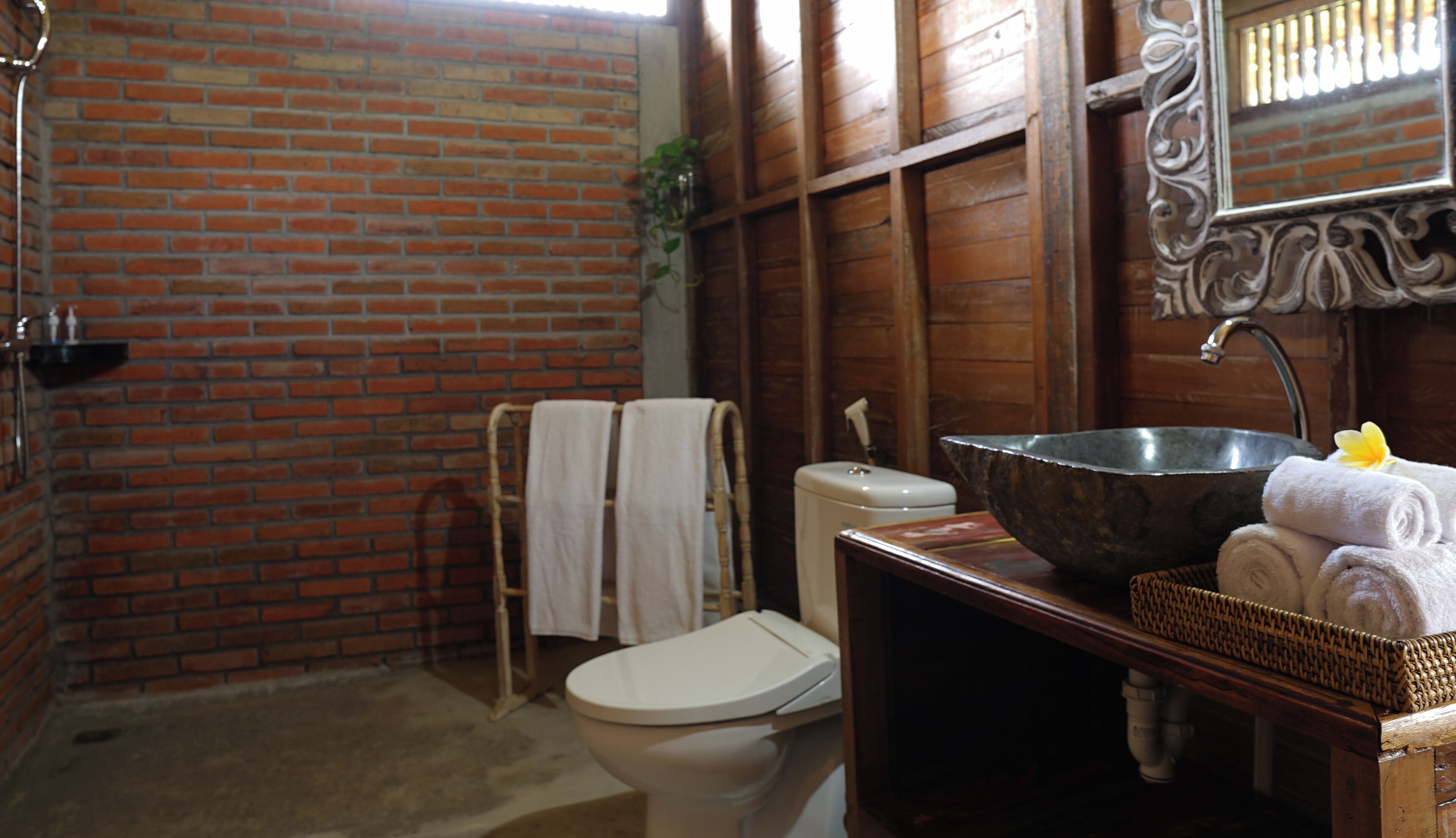 A bathroom with a brick wall, a white toilet, a large metal basin, and a wooden vanity.