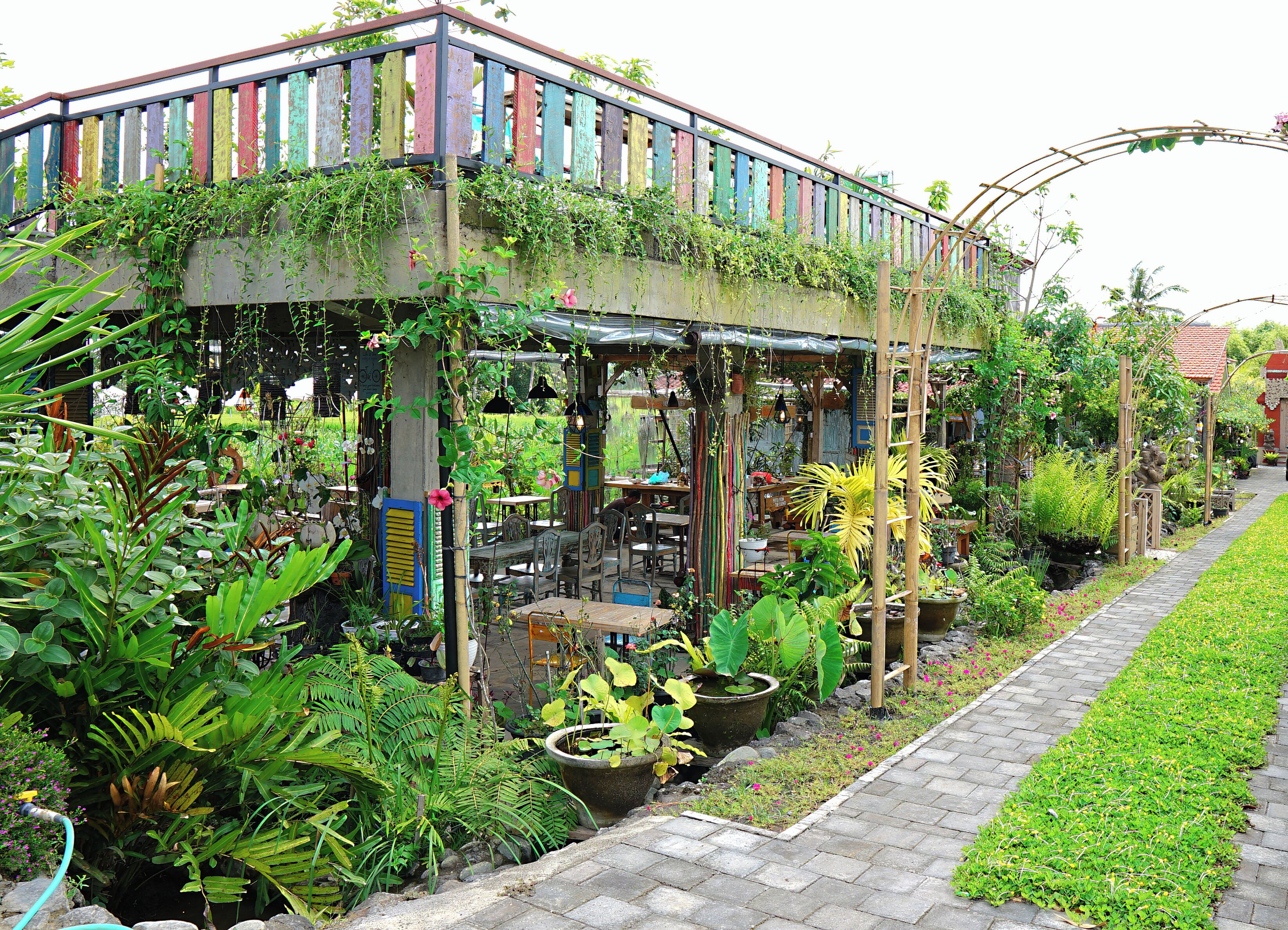 Cafetería en un jardín con mesas al aire libre, rodeada de abundante vegetación y diversas plantas.