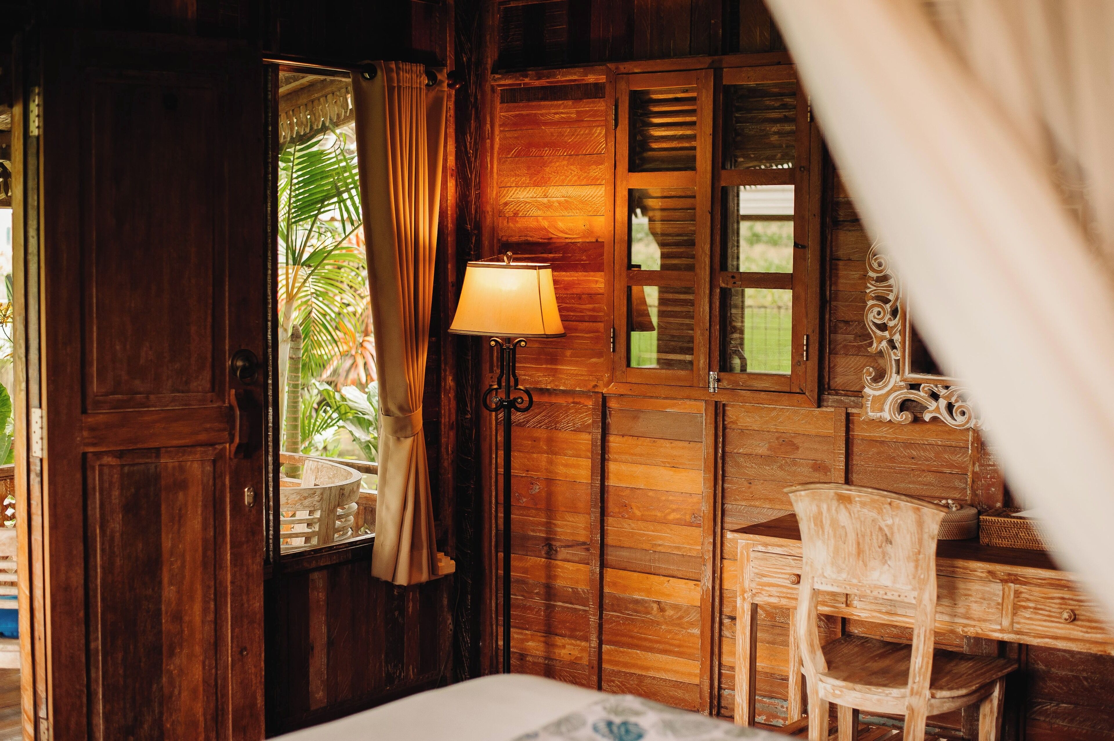 Ein Zimmer mit Holzboden, ausgestattet mit einem Bett, einem Schreibtisch und einem Stuhl. Ein Fenster mit Vorhängen und eine Lampe auf dem Schreibtisch.