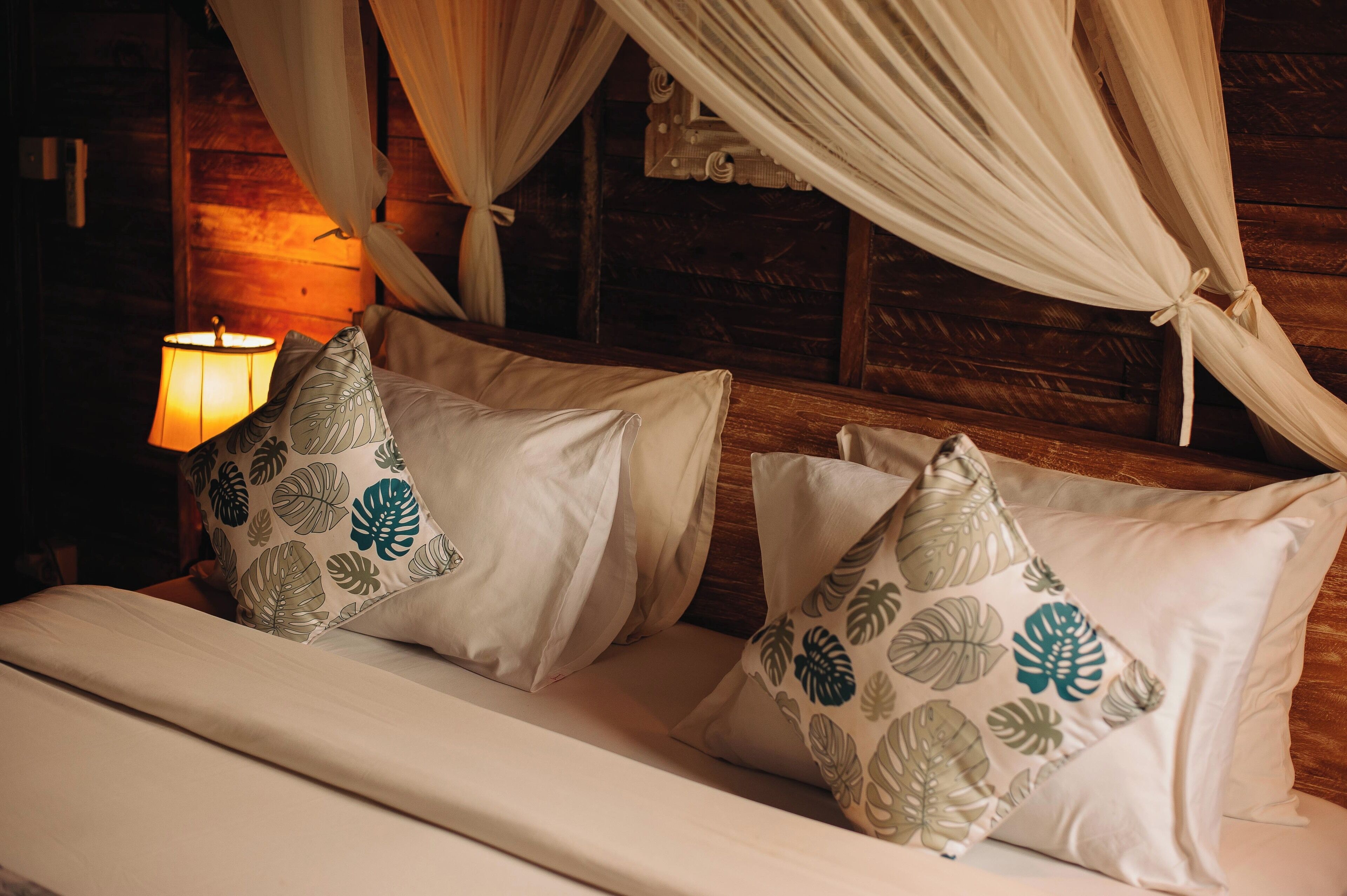Een bed met witte beddengoed en decoratieve kussens met bladmotieven, geplaatst tegen een houten wand met een hemelbed.