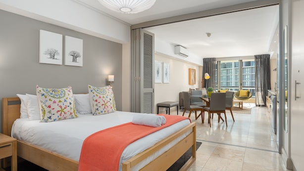 Ett modernt hotellrum med en säng i trä, färgglada kuddar, en röd pläd, en matplats med stolar och ett stort fönster med utsikt.
