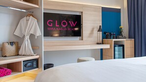 A modern hotel room with a bed, a TV mounted on the wall, a desk with a lamp, and a minibar. - GLOW Mira Karon Beach (Karon)