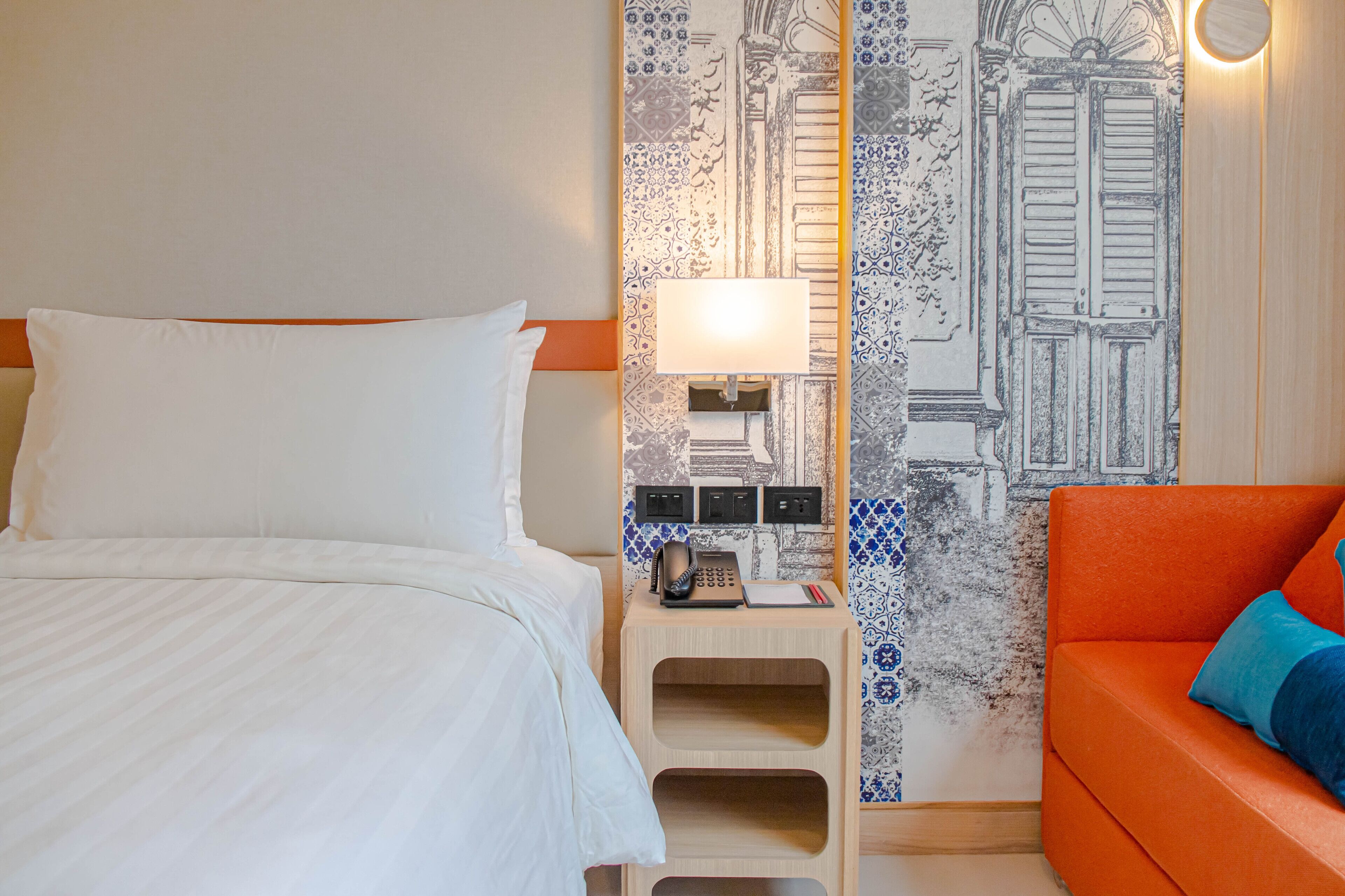 A hotel room with a bed, a nightstand with a lamp, a telephone, and a wall with a blue and white patterned design.