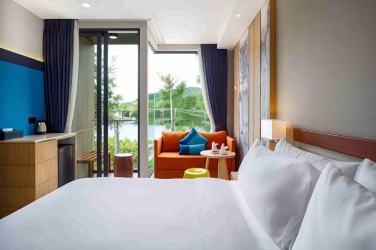 A hotel room with a large bed, a sofa, a small table, and a view of greenery outside.