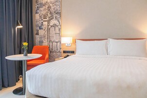 A hotel room with a large bed, a small table with a vase of flowers, an orange chair, a lamp, and a wall with a decorative pattern.