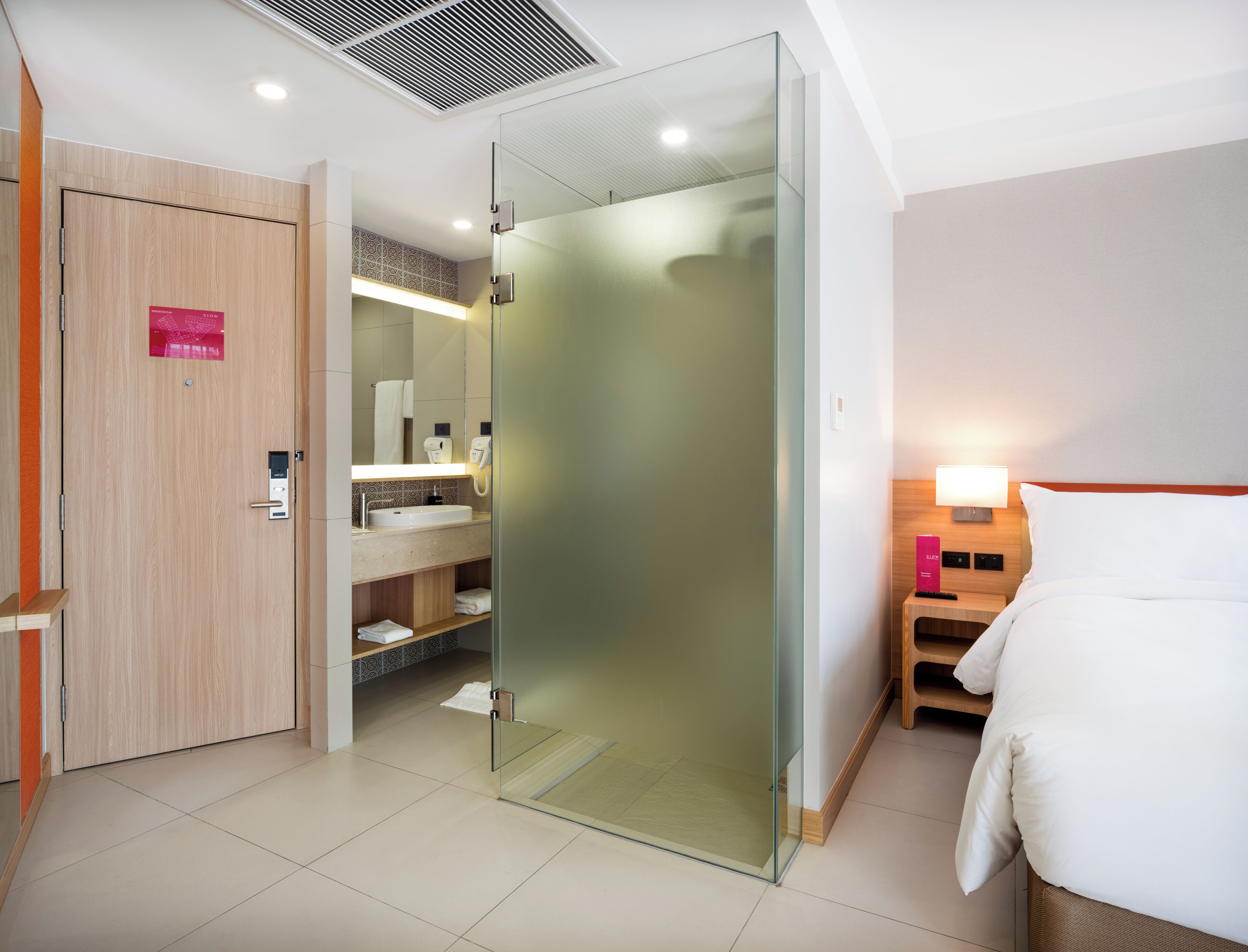 A modern hotel room with a glass shower enclosure, a wooden wardrobe, a bedside table with a lamp, and a bed with white bedding.