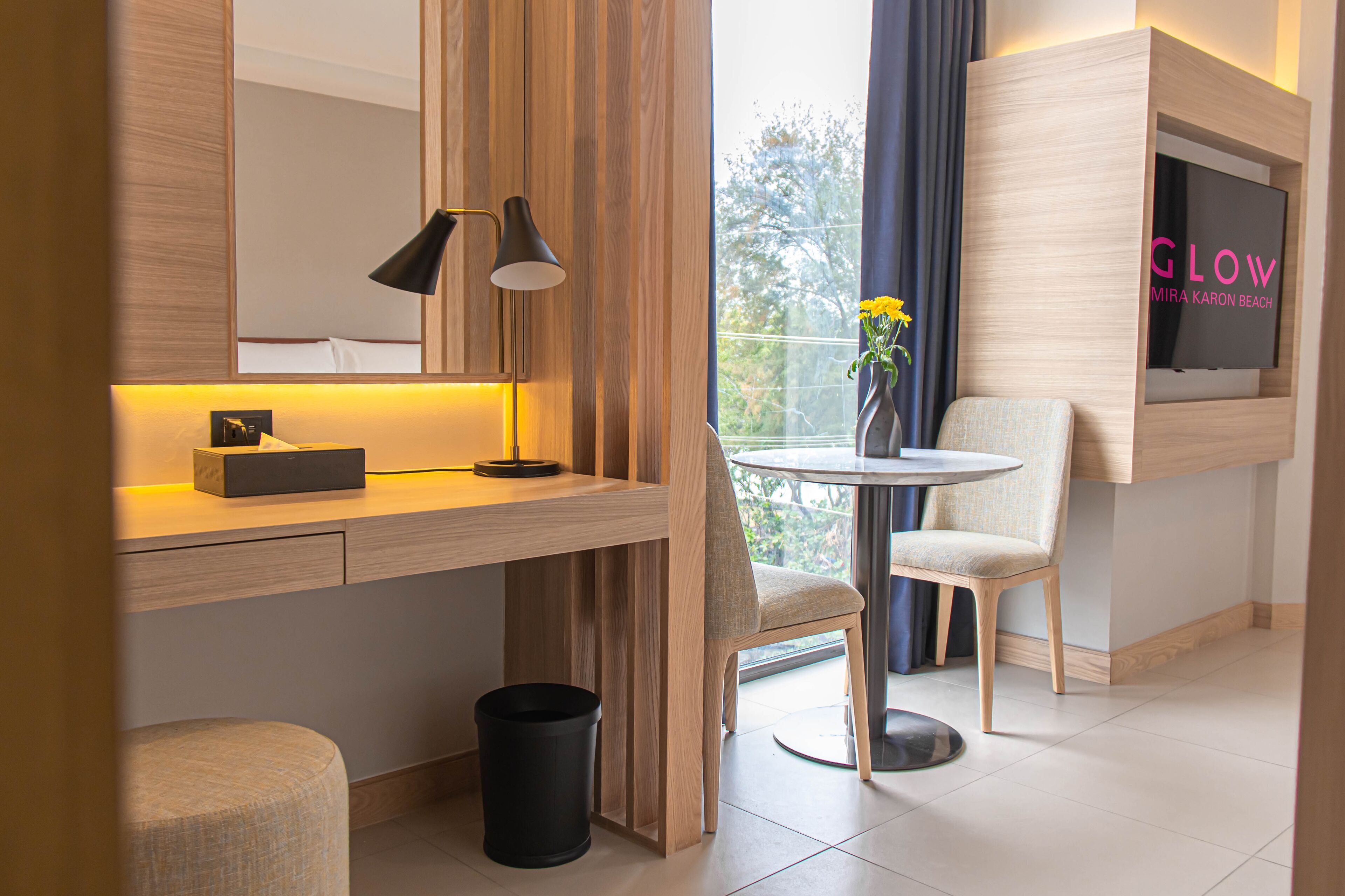 Una habitación de hotel moderna con un escritorio de madera, una mesita redonda pequeña, dos sillas, un televisor montado en la pared y una ventana con cortinas.