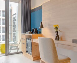 Una habitación de hotel moderna con un escritorio de madera, una silla, una cafetera, un jarrón con flores y vistas a la ciudad a través de amplias ventanas.