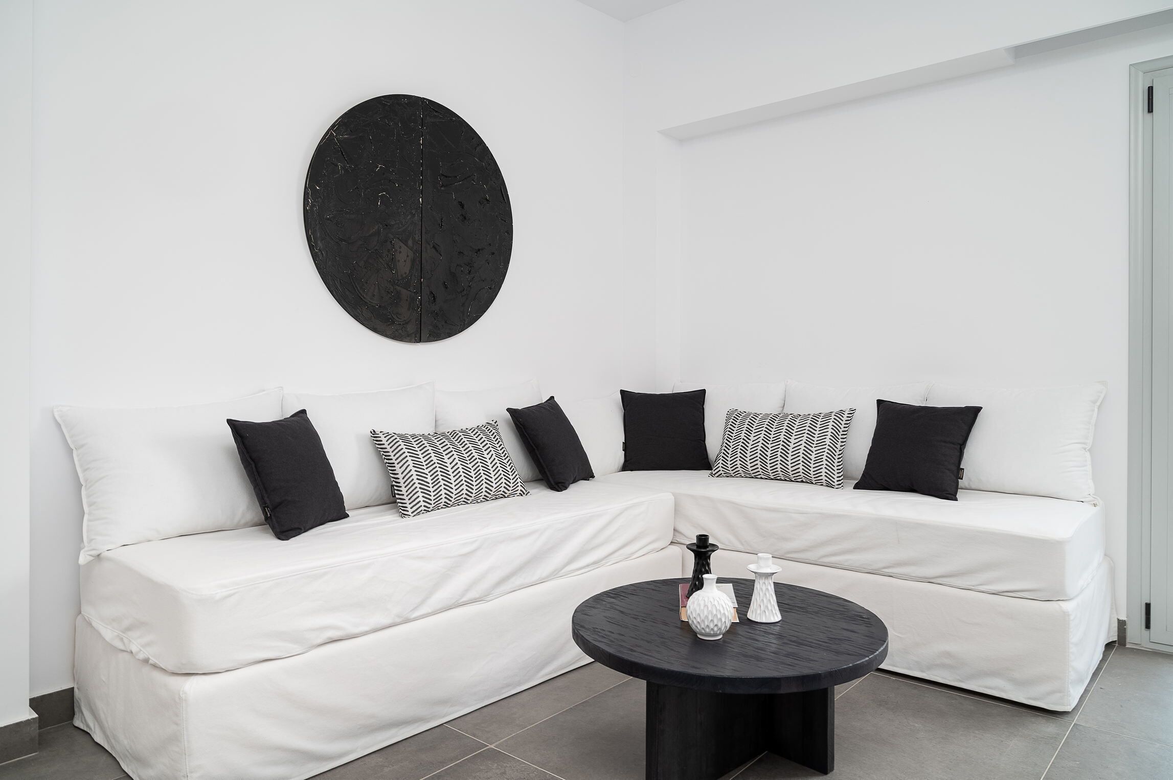 Un sofá modular blanco con cojines de estampado blanco y negro, una obra de arte circular negra en la pared y una mesa de centro negra con dos jarrones blancos.