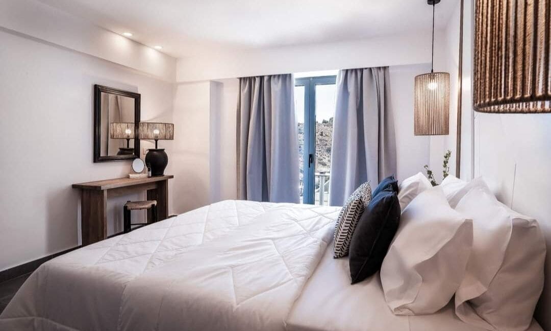 A hotel room with a large bed, a desk with a lamp, and a view of the outdoors through the window.