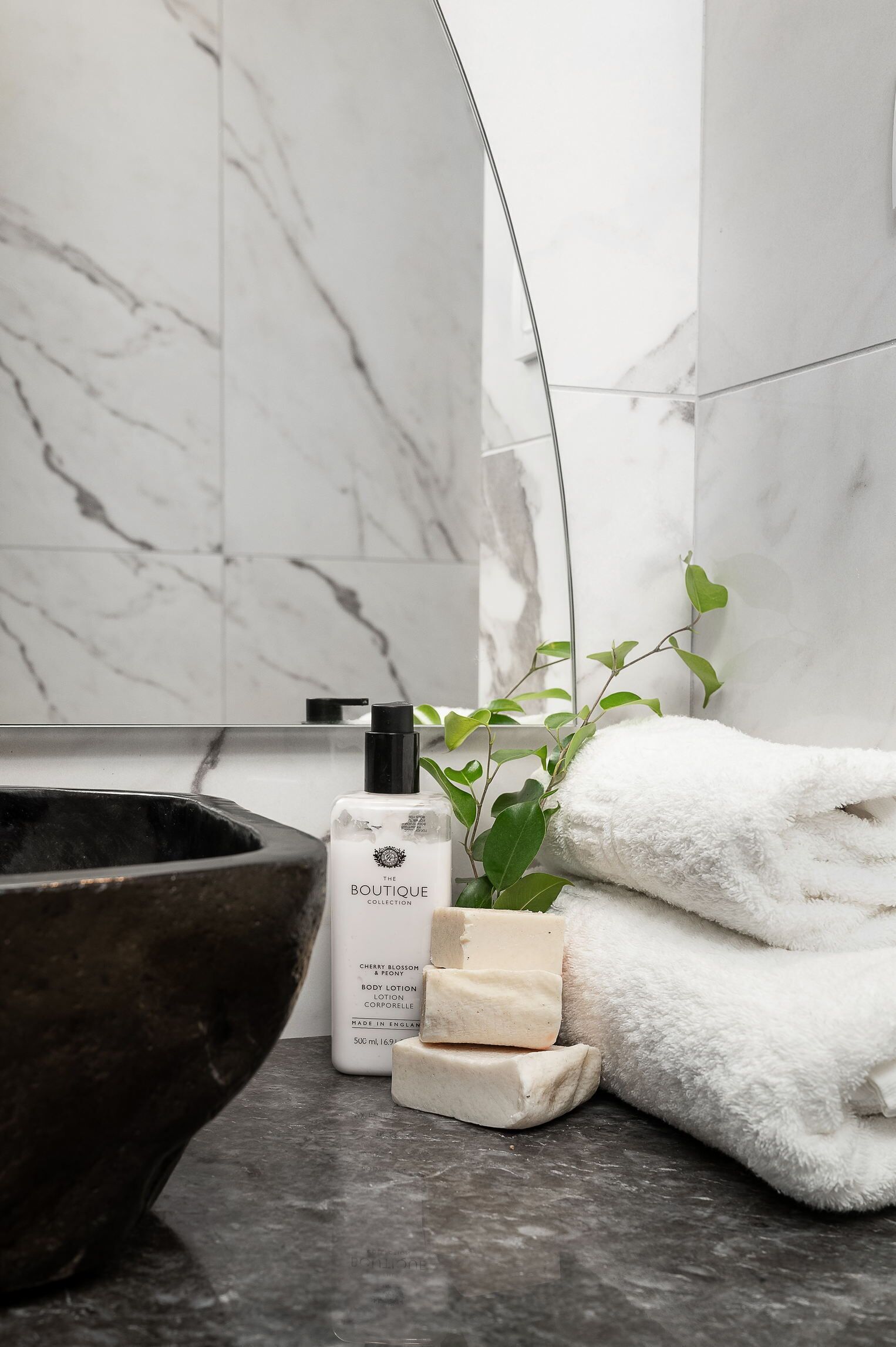 Une salle de bain aux murs de marbre, avec un lavabo noir, un flacon de lotion pour le corps et une serviette blanche.