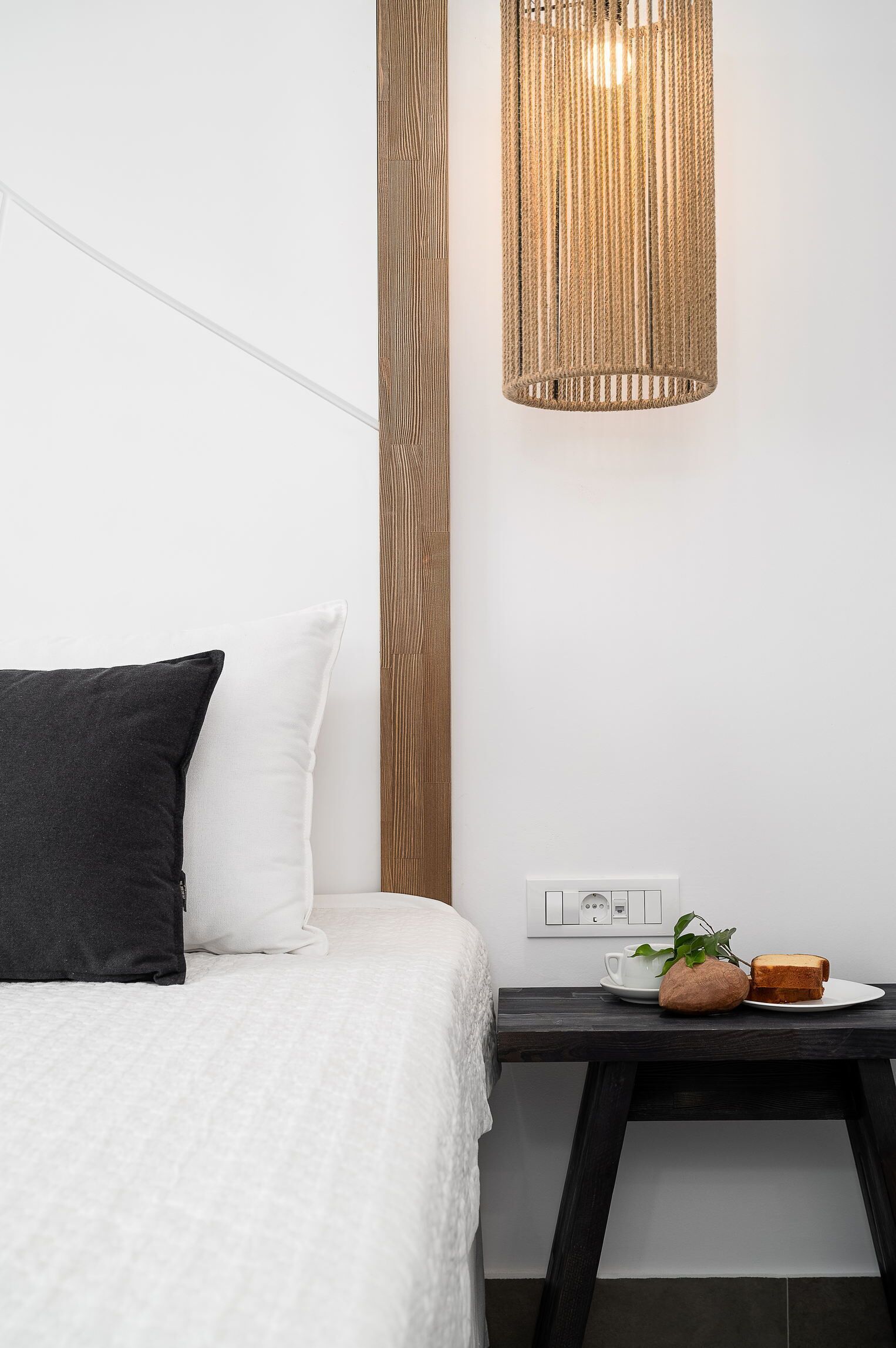 A neatly made bed with white and black pillows, a small bedside table with a plant, a cup, and a plate with bread.