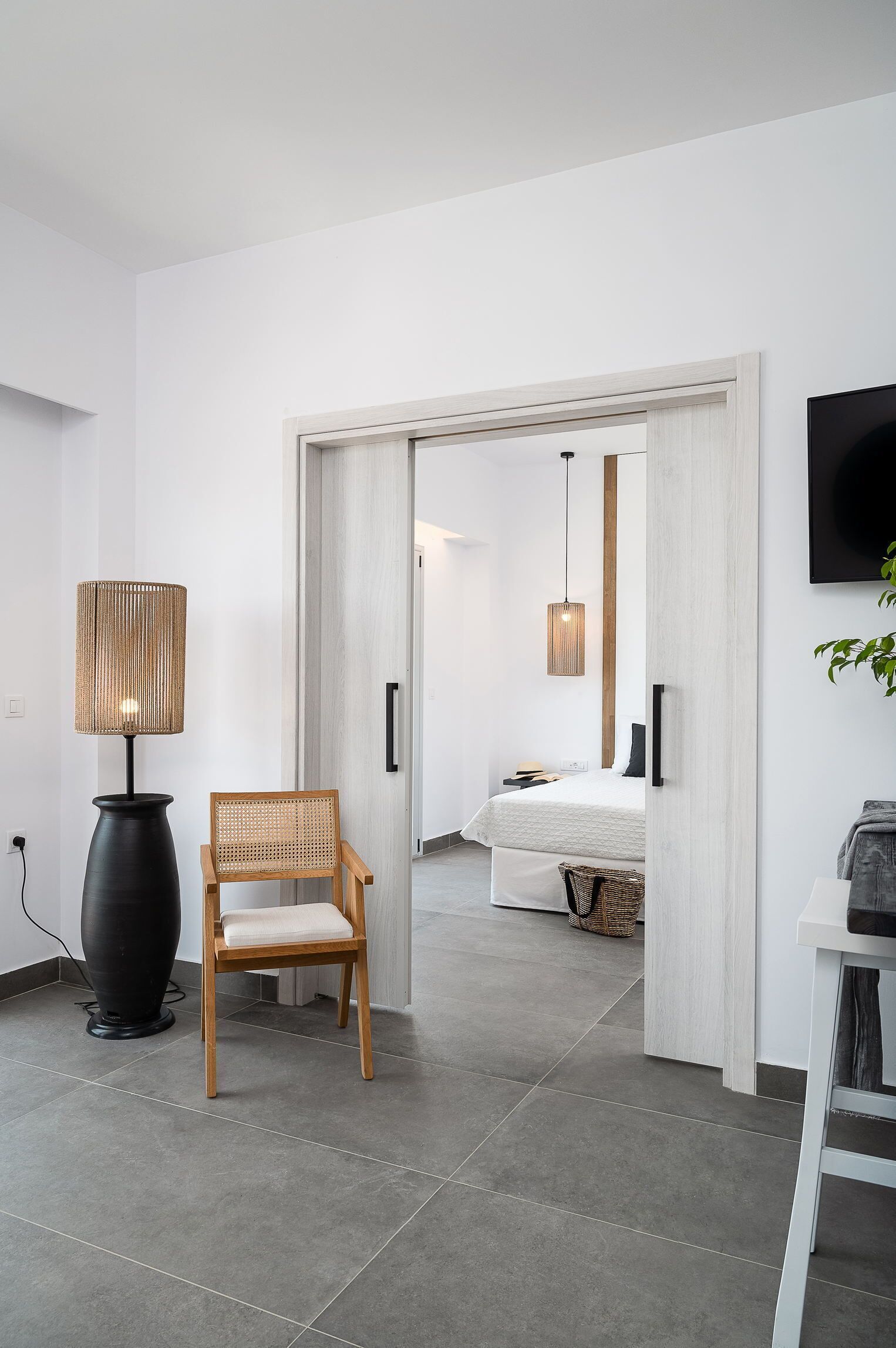 A modern hotel room with a large bed, a wooden chair, a floor lamp, and a TV on the wall.